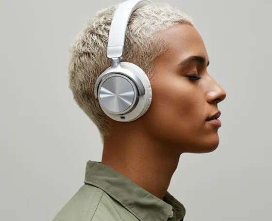 Profile of a person with short blonde hair wearing silver over-ear headphones and an olive green shirt against a neutral background.