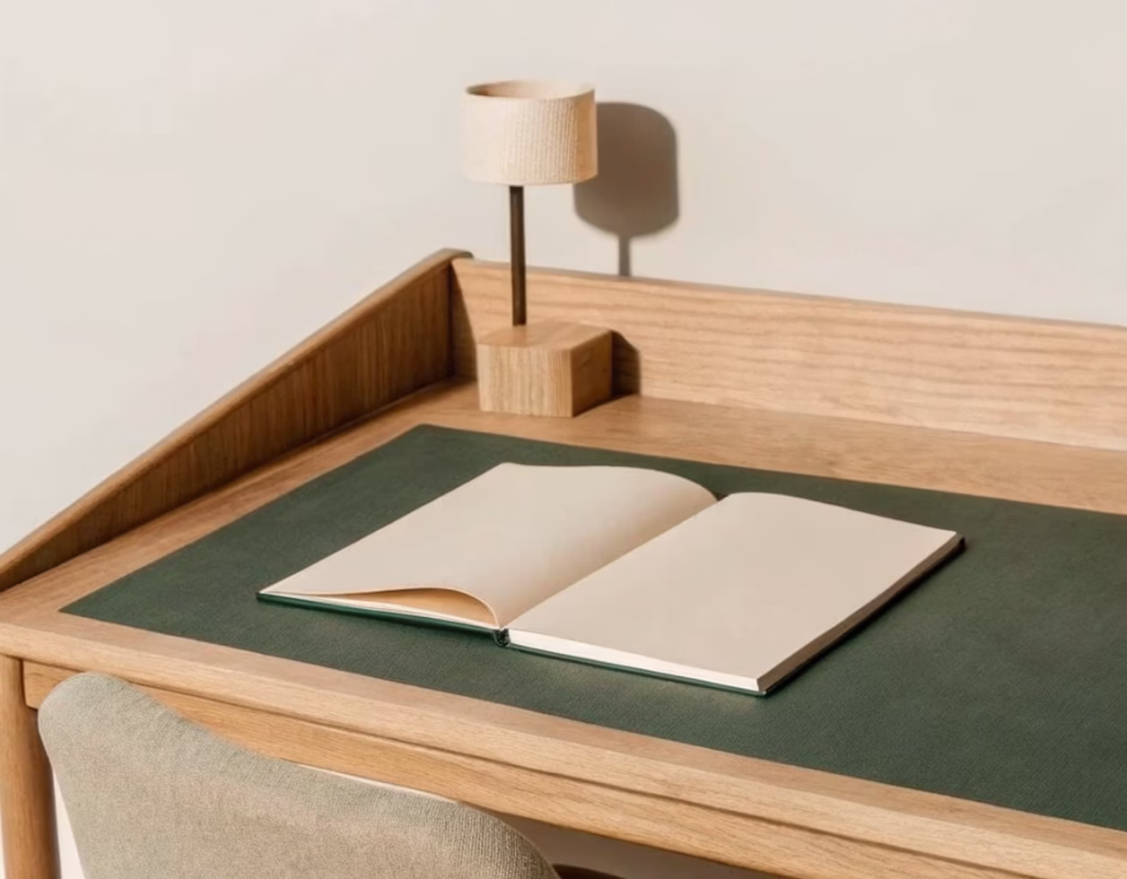 Minimal wooden desk with a green leather mat, an open blank book, and a small wooden lamp casting a shadow on the wall.