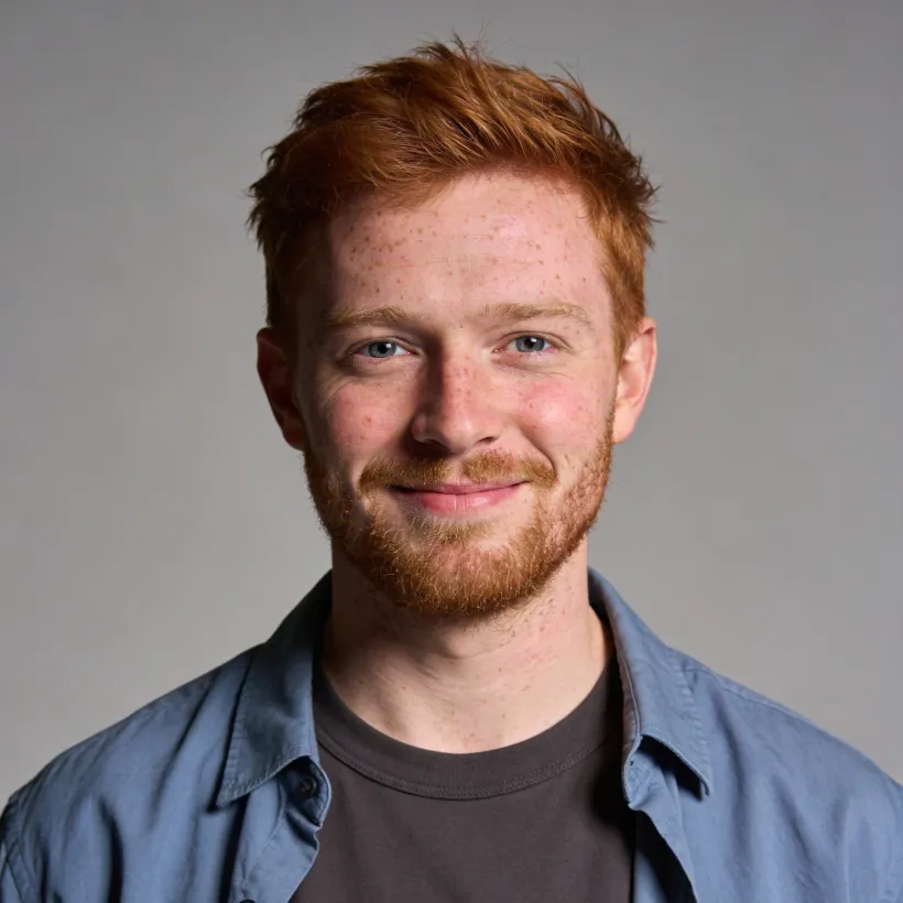Smiling young man with red hair and beard wearing a blue shirt over a dark t-shirt against a gray background.